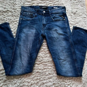 Replay Anbass - Säljer ett par Replay Anbass jeans i klassisk blå denim med snygg wash. De har fem fickor och snygga subtila slitningar. Jeansen har slim fit passform och raka ben, med detaljerade sömmar på bakfickorna och metallknapp framtill. Perfekta för dig som gillar stilrena jeans med lite edge. Jeansen är i storlek L29/W32 men passar också L28/W32. Perfekta för en stilren look på sommaren.