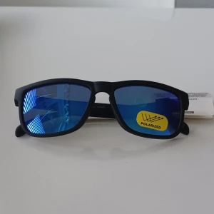 Svarta solglasögon med blåtonade polariserade linser från Foster Grant - Snygga svarta solglasögon från Foster Grant med rektangulär form och blåtonade polariserade linser. Bågarna är mattsvarta och tillverkade i plast. Perfekta för soliga dagar och ger 100% UVA-UVB skydd.