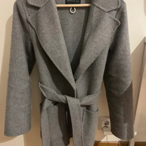 Grå kappa  - Snygg grå kappa med klassisk krage och bälte i midjan från Stockh LM. Kappan har lång ärm och en rak, stilren passform. Perfekt för kyligare dagar och enkel att matcha med olika outfits.