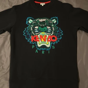 Svart sweatshirt med tigerbrodyr från Kenzo - Svart sweatshirt från Kenzo med ikonisk tigerbrodyr och färgglada detaljer framtill. Tröjan har rund halsringning och är tillverkad i mjuk bomull. Perfekt för dig som vill sticka ut med en statement-piece.