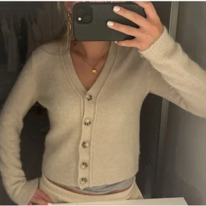 Beige croppad kofta med v-ringning - Säljer en beige croppad kofta med v-ringning och knappar framtill. Tröjan har långa ärmar och är i ett mjukt, stickat material. Perfekt att styla med både jeans och kjol.