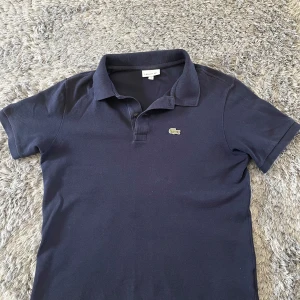 Mörkblå piké t-shirt från Lacoste - Säljer en klassisk mörkblå piké t-shirt från Lacoste med den ikoniska krokodilloggan broderad på bröstet. T-shirten har krage och korta ärmar, perfekt för en stilren look.