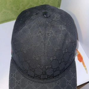 Svart keps från Gucci med monogram - Säljer en svart keps från Gucci med diskret GG-monogram över hela kepsen. Kepsen har en klassisk form, justerbar rem i skinn baktill och en grön-röd rand på sidan. Perfekt accessoar för en stilren look.