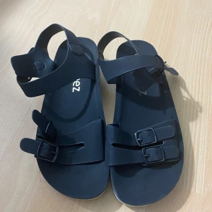 Svarta sandaler från Beez - Svarta sandaler från Beez med tre justerbara remmar och spännen. Snygg och enkel design med platt sula, perfekt för varma dagar. Tillverkade i syntetmaterial.