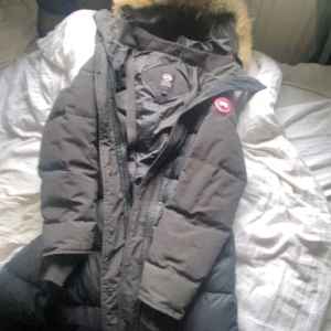Svart dunjacka från Canada Goose med pälskant - Säljer en svart dunjacka från Canada Goose med stor huva och pälskant. Jackan har lång ärm, dragkedja och knappar framtill samt klassisk logga på ärmen. Perfekt för kalla dagar med sin varma och fluffiga design. Då den inte kommer till användning längre är i perfekt schik storlek M men passar både L och S använd 2gånger pris kan diskuteras 