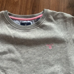 Grå sweatshirt från Gant med rosa detaljer - Grå sweatshirt från Gant med rund halsringning och diskret rosa logga broderad på bröstet. Mjukt material och rosa insida vid halsen. Perfekt för en avslappnad klädsel! 💓