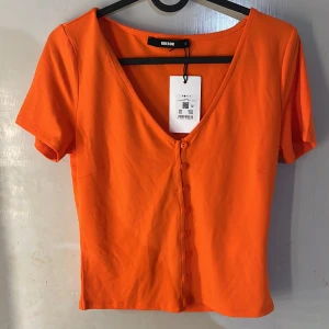 Topp  - Säljer en orange topp från BikBok med v-ringning och knappar framtill. Toppen har korta ärmar och är tillverkad i mjuk bomull. Perfekt för dig som gillar färgstarka plagg och enkel stil.