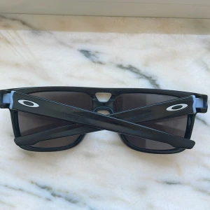 Svarta solglasögon från Oakley - Nypris 1995!     Snygga svarta solglasögon från Oakley med rektangulära linser och diskret logga på skalmarna. Glasögonen har en sportig design och passar perfekt för soliga dagar.