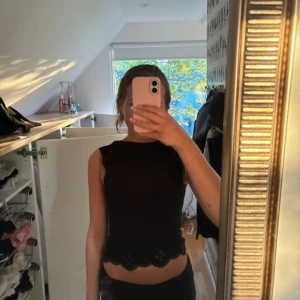 Svart croppad topp med spets från Zara - Säljer en svart croppad topp från Zara med fina spetsdetaljer och blommönster längs nederkanten. Toppen är ärmlös och har en tajt passform. Toppen kan bäras på två sätt