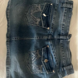 Lågmidjad Jeanskjol med paljettfickor - Snygg lågmidjadblå jeanskjol med två bakfickor dekorerade med paljetter. Kjolen har bälteshällor och dragkedja framtill. Perfekt för dig som gillar denim med lite extra detaljer. Sitter  som s 
