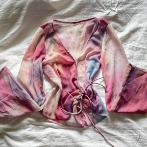 Omlottopp i pastellfärger - Säljer en omlottopp med långa, vida ärmar i tunnt, transparent material. Toppen har ett vackert mönster i rosa, lila och blå toner och knyts i midjan. Perfekt för dig som gillar unika och färgglada plagg.