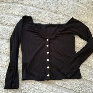 Svart ribbad topp med knappar - Svart långärmad topp med ribbad struktur och vita knappar framtill. Djup v-ringning och croppad passform, perfekt för en trendig look. Enkel att matcha med både jeans och kjol.