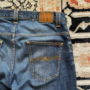 Mörkblå jeans från Nudie Jeans - Säljer ett par mörkblå jeans från Nudie Jeans med klassiska orange sömmar och snygga detaljer på bakfickorna. Jeansen har en rak passform och är tillverkade i slitstarkt denim. Perfekta för dig som gillar stilrena och tidlösa jeans. Skick 10/10. Pris 499. Tveka inte att höra av dig!