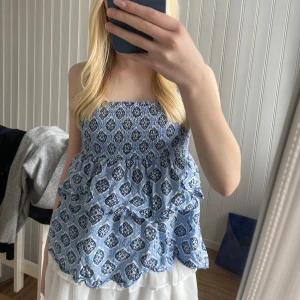 Blåmönstrad smockad linnetopp - Säljer en blå och vit linnetopp med smockad överdel och volanger. Toppen har tunna justerbara axelband. Den är knövlig på bilderna men det är bara för att den är ostruken!