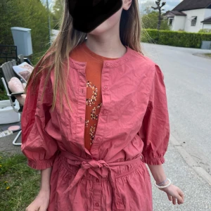 Rosa klänning med knytband i midjan - Säljer en söt rosa klänning med trekvartsärmar och knytband i midjan. Klänningen har rund halsringning och knappar framtill. Perfekt för vår och sommar!