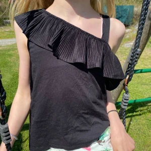 Svart oneshoulder topp med volang - Ett supergulliga linne som knappast är använt! Personen på bilden är ca 140 cm och linnet passar lite olika storlekar! Titta gärna på min Vinted ” estridgavmigtips ” där denna och många fler annonser är publicerade🌸