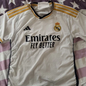 Real Madrid fotbollströja Adidas - Vit Real Madrid fotbollströja från Adidas med svarta och gula detaljer. Tröjan har korta ärmar med gula ränder på axlarna och klubbmärke på bröstet. Svarta sidopaneler och sponsortryck på framsidan. Storlek L men passar också M