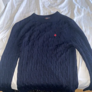 Mörkblå stickad tröja från Morris - Mörkblå stickad tröja från Morris i 100% merinoull. Klassisk modell med rund halsringning och diskret röd logga broderad på bröstet. Pris kan diskuteras