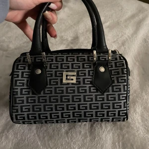 Svart och grå handväska  - Säljer en liten och stilren handväska med svart och grått mönster. Väskan har svarta handtag och silverfärgade metalldetaljer. Perfekt accessoar för att lyfta din outfit.