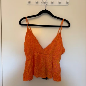 Orange mönstrad linnetopp - Säljer en orange linnetopp med smala axelband och v-ringning. Toppen har ett blommigt mönster i rosa och en lätt volang nedtill. Perfekt för varma dagar och somriga outfits.