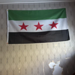 Syriska frihetsflaggan - Säljer en syrisk frihetsflagga med tre horisontella ränder i grönt, vitt och svart samt tre röda stjärnor i mitten. Flaggan är rektangulär och tillverkad i tyg, perfekt att hänga på väggen.