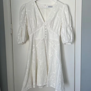 Vit blommig klänning med puffärm från Adoore - Superfin vit klänning från Adoore med v-ringning, puffärmar och knappar framtill. Klänningen har broderade blommor och markerad midja. Perfekt för sommarens alla tillfällen!💕