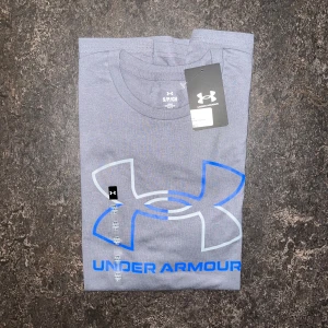 Grå t-shirt från Under Armour - Snygg grå t-shirt från Under Armour med stor logga i blått och vitt framtill. Klassisk rund halsringning och korta ärmar. Perfekt för träning eller chill dagar. Materialet känns mjukt och skönt mot huden.