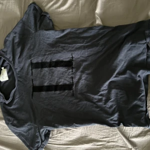 Mörkblå t-shirt från Les Deux - Säljer en mörkblå t-shirt från Les Deux med två svarta broderade ränder på bröstet. Klassisk passform och korta ärmar. Perfekt för en stilren och enkel look.