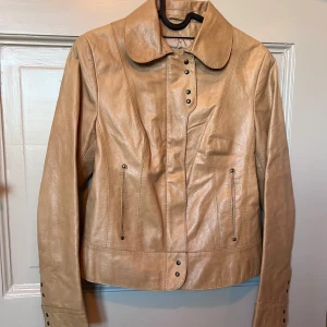 Beige äkta skinnjacka med knappar - Säljer en beige skinnjacka med snygga detaljer och stora knappar framtill. Jackan har lång ärm, markerad krage och två fickor med dragkedja. Äkta skinn / läder. Storlek S.