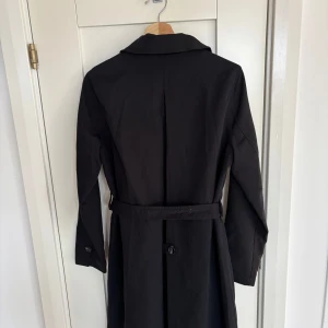 Svart trenchcoat från Happy Holly - Snygg svart trenchcoat från Happy Holly med knappar framtill och bälte i midjan. Klassisk krage och långa ärmar. Yttermaterial i 65% polyester och 35% bomull, fodrad med randigt tyg. Perfekt för dig som vill ha en stilren och tidlös jacka.