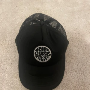Svart keps från Rip Curl - Säljer en svart keps från Rip Curl med rund vit logotyp framtill. Kepsen har nät baktill för extra ventilation och justerbar passform. Perfekt för en avslappnad stil.