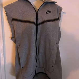 Grå ärmlös kofta nike tech - Snygg grå kofta från Nike med dragkedja framtill och huva. Västen har svarta detaljer och två fickor på sidorna. Perfekt för lager-på-lager och sportig stil. Gjort en egen liten design på denna kofta, har klippt av båda armarna men den funkar att ha ändå. Väldigt fin om du frågar mig.