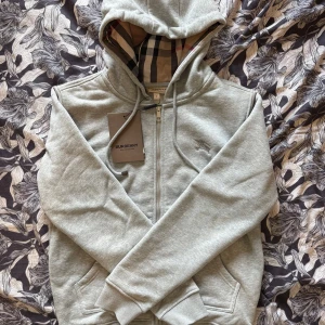 HELT NY BURBERRY ZIP-UP - Säljer en grå hoodie från Burberry med klassiskt rutigt foder i huvan. Jackan har dragkedja framtill, fickor och ribbade muddar. Perfekt för en stilren och avslappnad look.