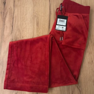 Röda mjukisbyxor med fickor juicy couture  - Snygga röda mjukisbyxor från Juicy Couture i en mjuk och skön velour. Byxorna har praktiska fickor med knappar och en justerbar dragsko i midjan. Perfekta för en avslappnad stil med en touch av lyx.