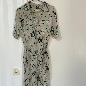 Blommig klänning  - Snygg blommig klänning från vero Moda i en ljusgrön nyans med rosa och svarta blommor. Klänningen har korta ärmar och ett knytband i midjan för en smickrande passform. Perfekt för vår och sommar!