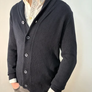 Svart kofta med knappar - Snygg svart kofta med knappar framtill. Den har en klassisk design med ribbade muddar och en v-ringad krage. Perfekt för lager-på-lager-stil. Inga defekter, bra skick. Modellen är 182cm/67kg