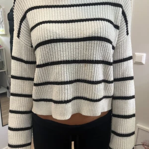 Vit och svart randig stickad tröja från Kappahl - Säljer en snygg vit stickad tröja från Kappahl med svarta ränder. Tröjan har en croppad passform och långa ärmar, perfekt för en stilren look. Den är både bekväm och trendig, idealisk för kyligare dagar.