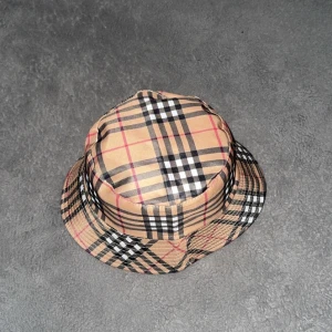 Rutig bucket hat från Burberry  - Snygg bucket hat med klassiskt rutigt mönster i beige, svart, vit och röd. Perfekt för att ge din outfit en stilren touch. Passar bra till både vardag och speciella tillfällen.