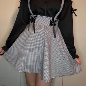 Svartvit rutig kjol med hängslen - Charmig svartvit rutig kjol med korsade hängslen och spetsdetaljer. Kjolen har dekorativa rosetter och små korsdetaljer framtill. Perfekt för klassisk jirai kei 