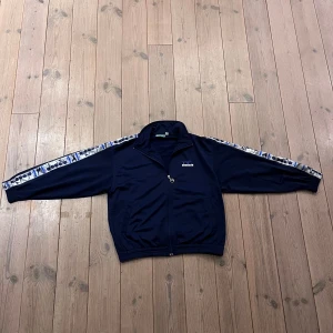 Vintage träningsjacka från Diadora - Säljer en snygg mörkblå träningsjacka från Diadora med dragkedja och mönstrade detaljer längs ärmarna. Perfekt för en sportig look. Jackan har en normal passform och är långärmad. 