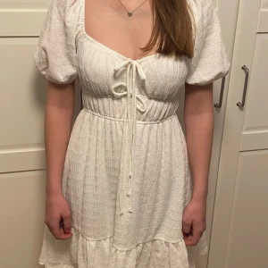 Vit klänning från Gina Tricot - Charmig vit klänning från Gina Tricot med puffärmar och knytning framtill. Den har en volangdetalj vid fållen och en smickrande passform. Perfekt för sommarens alla tillfällen!