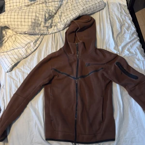 Brun huvtröja från Nike - Säljer en brun huvtröja från Nike med dragkedja och en ficka på ärmen. Perfekt för kyliga dagar med sin långa ärmar och bekväma passform. Huvan ger extra skydd mot väder och vind.
