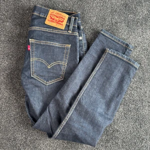 Slim Levis jeans  - Säljer ett par klassiska Slim Levis jeans i mörkblå denim med kontrasterande sömmar. De har en traditionell femficksdesign och en läderpatch med Levis logotyp på baksidan. Perfekta för en tidlös stil.