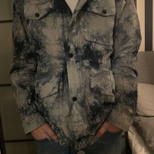 Tie Dye Hiking Jacka från J. Lindeberg - Nypris 6000. Helt oanvänd. Jackan ser ut att vara vit och blå på bild men är grön och blå i verkligheten.  Säljer en cool tie dye hiking jacka från J. Lindeberg i färgen grön och blå.  Jackan har en unik mönsterdesign och är perfekt för utomhusaktiviteter. Den har långa ärmar och praktiska fickor framtill. 