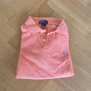 Korallfärgad pikétröja från Ralph Lauren - Säljer en snygg korallfärgad pikétröja från Ralph Lauren. Tröjan har en klassisk krage med två knappar och det ikoniska broderade logotypen i blått på bröstet. Perfekt för en stilren och avslappnad look.