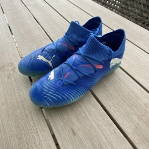 Blå fotbollsskor från Puma - Snygga blå fotbollsskor från Puma med vit logga och grön sula. Skorna har snörning och en strumpliknande passform för extra komfort. Perfekta för fotbollsplanen med sin stilrena design och funktionalitet, Storlek 40. 