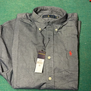 Grå/blå skjorta från Ralph Lauren - Snygg grå skjorta från Ralph Lauren med klassisk krage och knappar framtill. Den har en broderad logotyp i rött på bröstet och är långärmad. Perfekt för en stilren look.