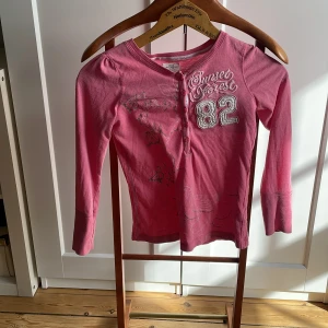 Rosa långärmad topp från L.O.G.G. - Säljer en rosa långärmad topp från L.O.G.G. med trycket 'Sunset Forest 82' på framsidan. Toppen har knappar vid halsringningen och ett blommigt mönster på ena sidan. 
