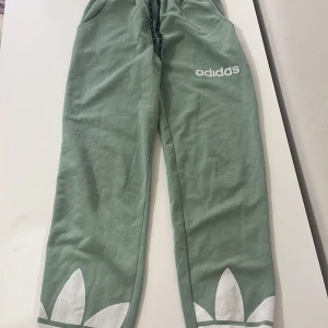 Gröna mjukisbyxor från Adidas - Snygga gröna mjukisbyxor från Adidas med elastisk midja och dragsko. Byxorna har vita detaljer och Adidas-logga på benet. Perfekta för en avslappnad stil.