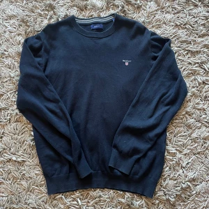 Svart tröja från GANT - Säljer en stilren svart tröja från GANT med rund hals och diskret logga på bröstet. Perfekt för en klassisk look. Tröjan har långa ärmar och ribbade muddar vid ärmslut och nederkant.